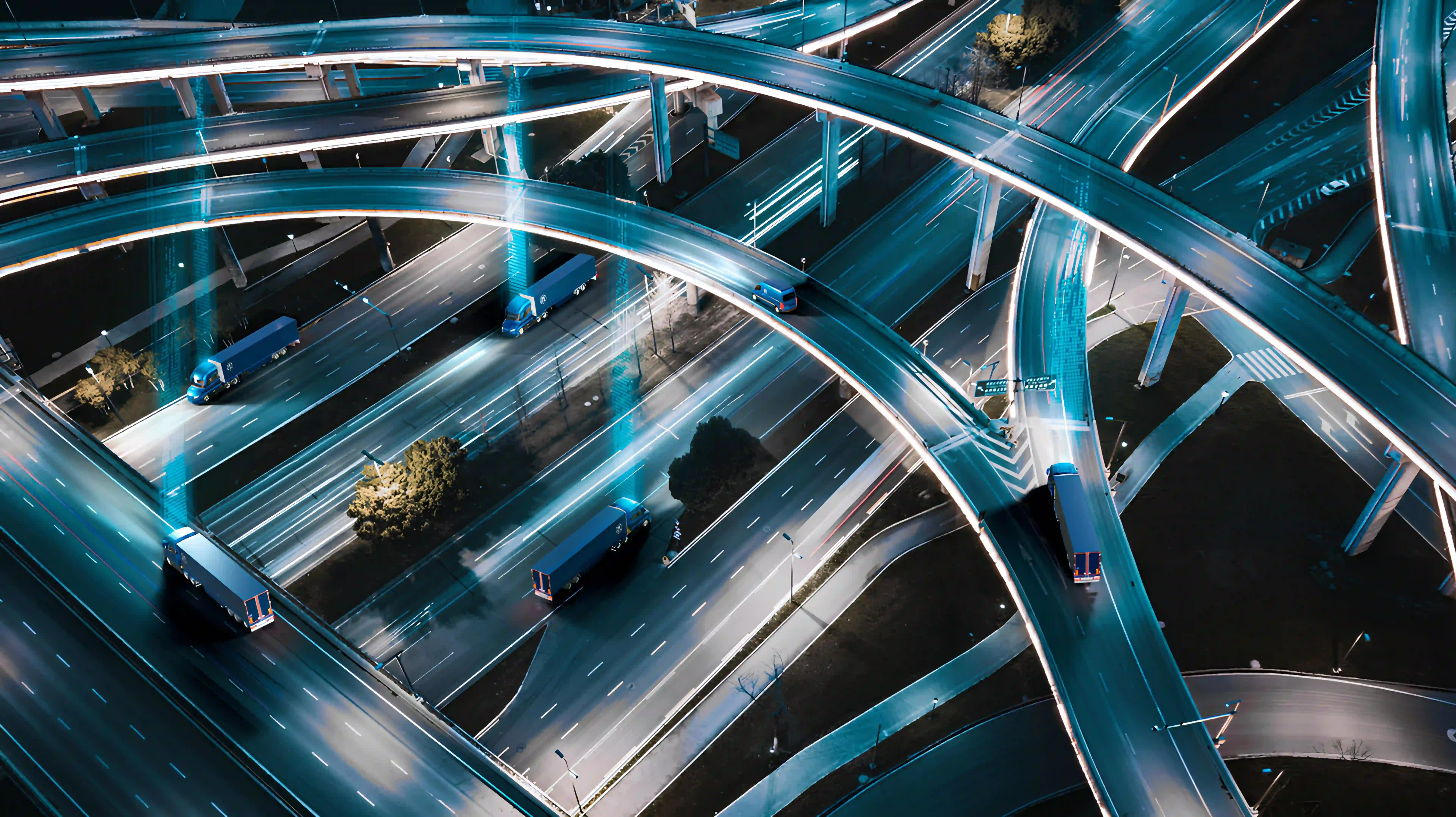 Aerial view of highway interchange at night with trailers on multiple routes — cyan light trails showing fleet movement as a connected network